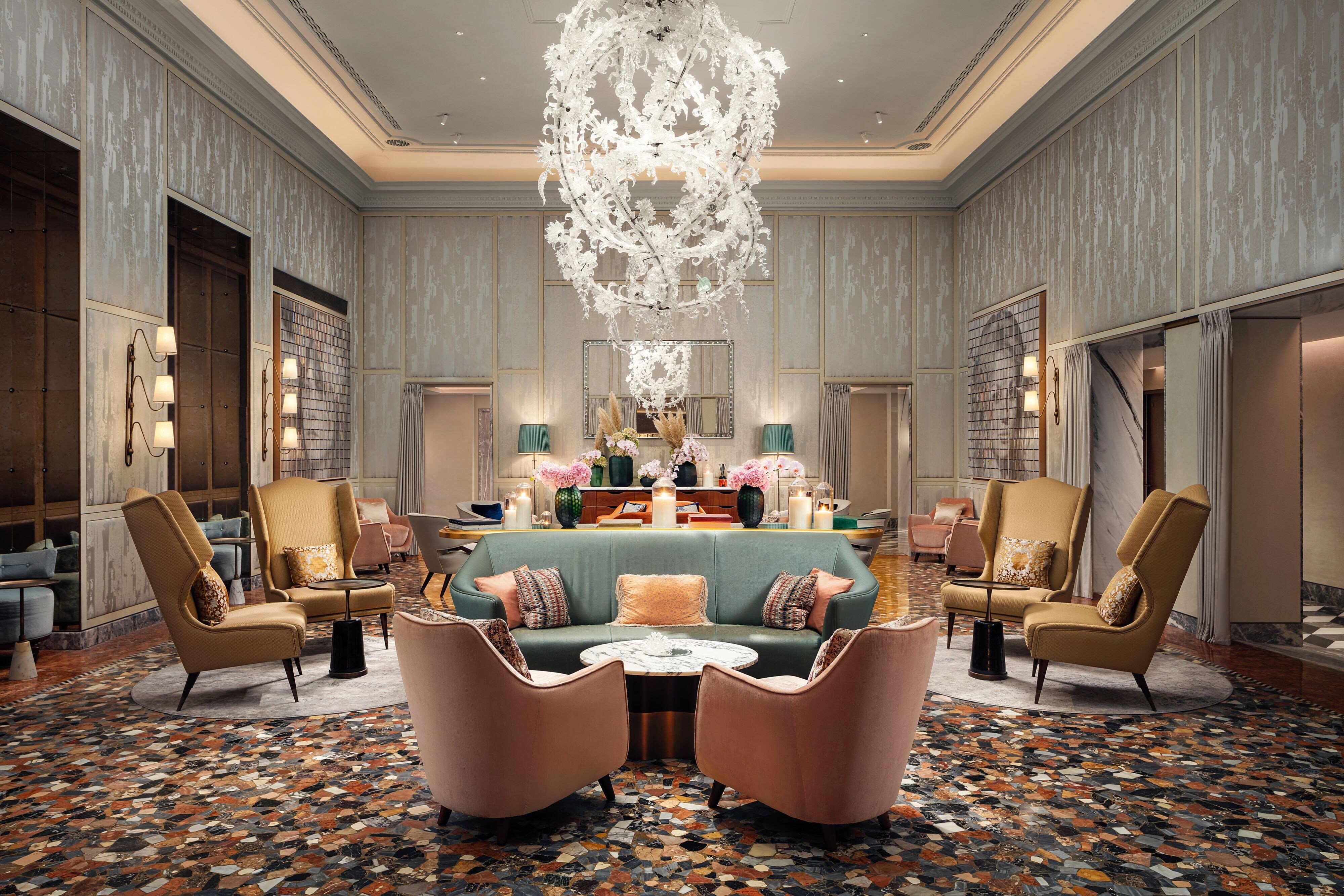The lobby lounge at The St. Regis Venice with a large Murano glass chandelier, Venetian terrazzo floor, a curved green sofa flanked by gold and pink armchairs, and candles on a central console