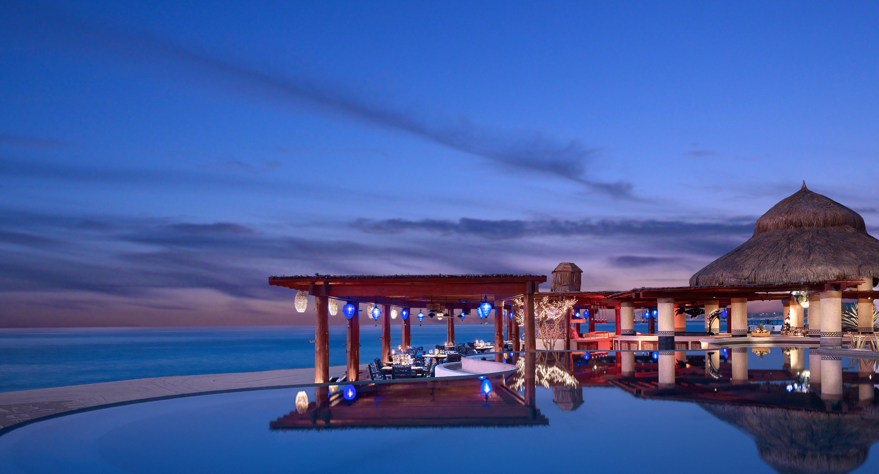A palapa-roofed restaurant pavilion lit with blue lanterns at dusk, reflected in a still infinity pool that runs to the Sea of Cortes at Las Ventanas al Paraiso