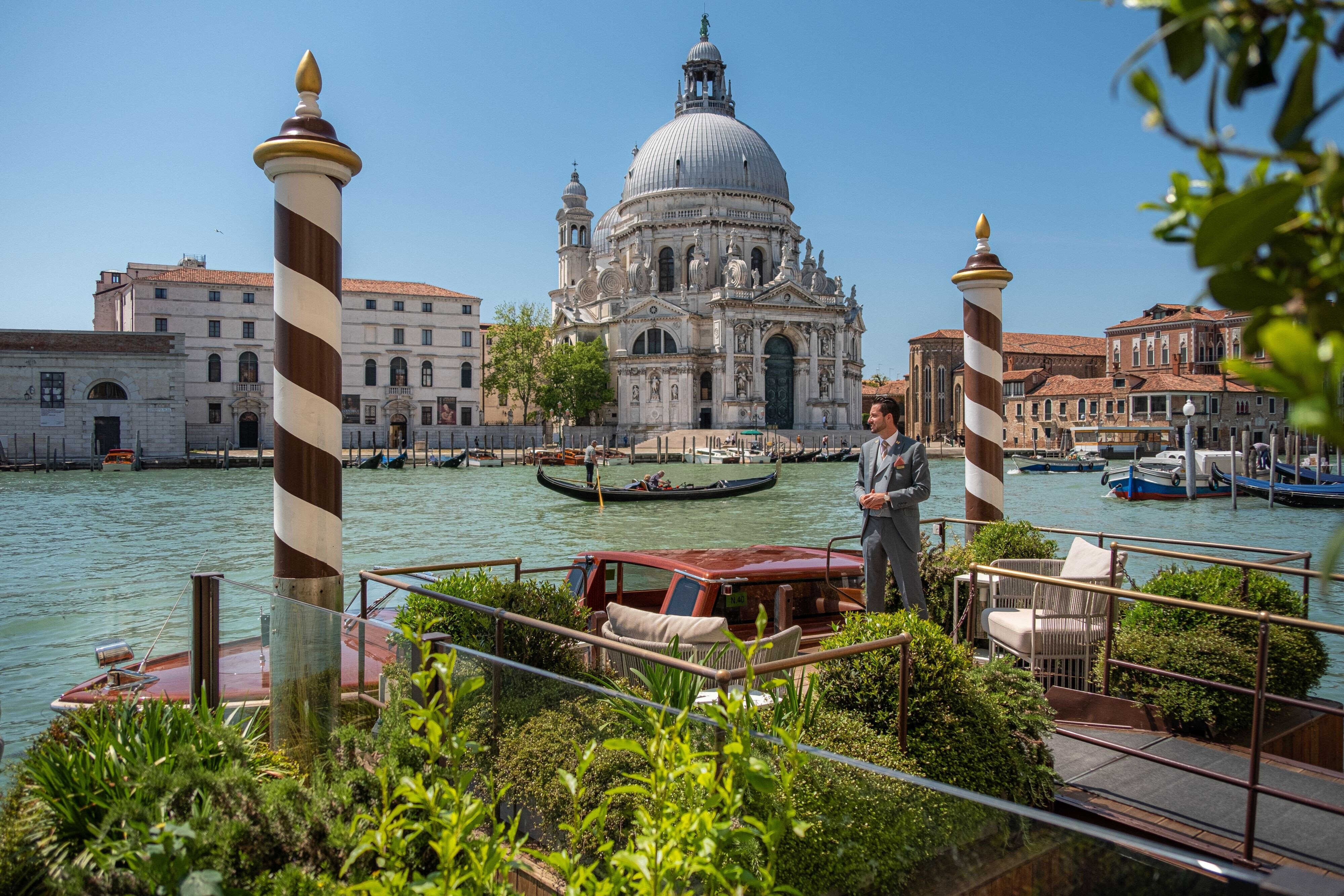 A St. Regis butler in a grey suit waiting at the private water entrance of The St. Regis Venice with a mahogany water taxi and the basilica of Santa Maria della Salute across the Grand Canal - the staffed service layer that inspection ratings score and star plaques only imply