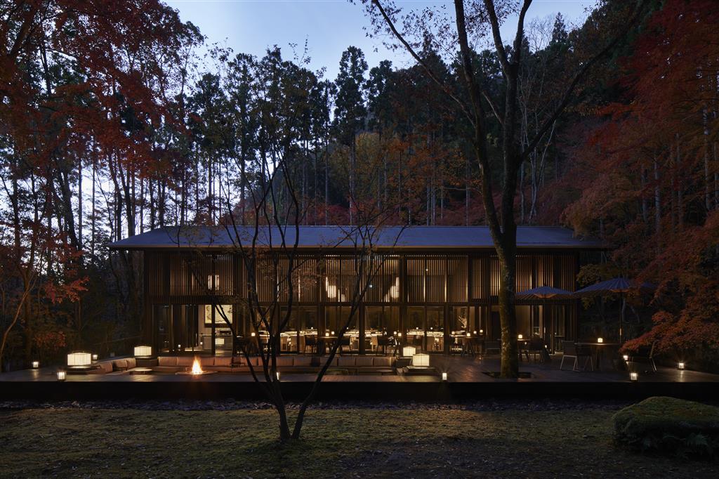 A low timber-and-glass pavilion lit from within at dusk at Aman Kyoto, its dining room visible through slatted screens, with a sunken fire pit and stone lanterns on the deck in front and red autumn maples and cedar forest rising behind