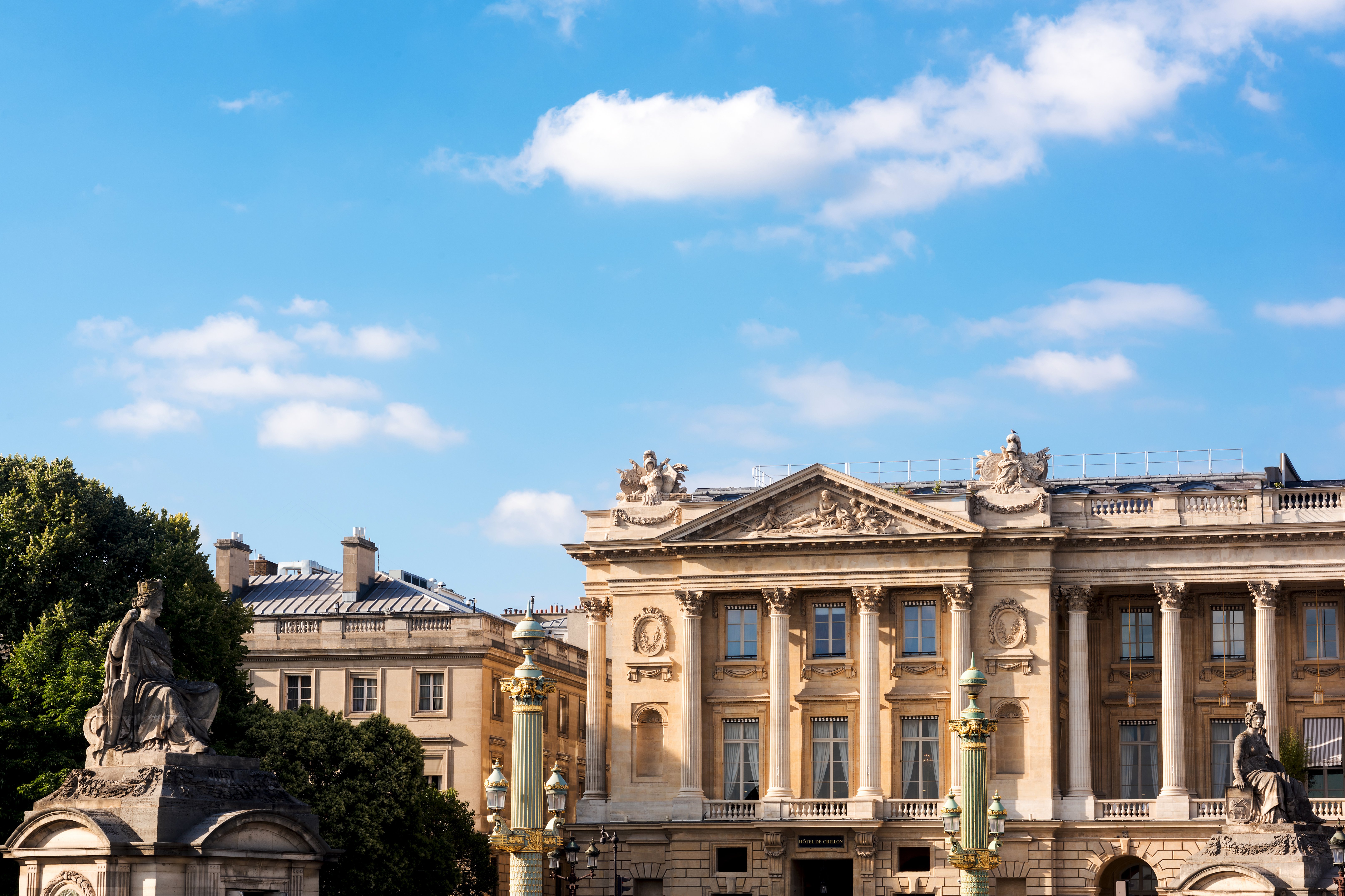 The colonnaded eighteenth-century facade of Hotel de Crillon on the Place de la Concorde in Paris, with the sculpted pediment, a green-and-gold Concorde lamp column and a seated stone statue in the foreground under a blue sky