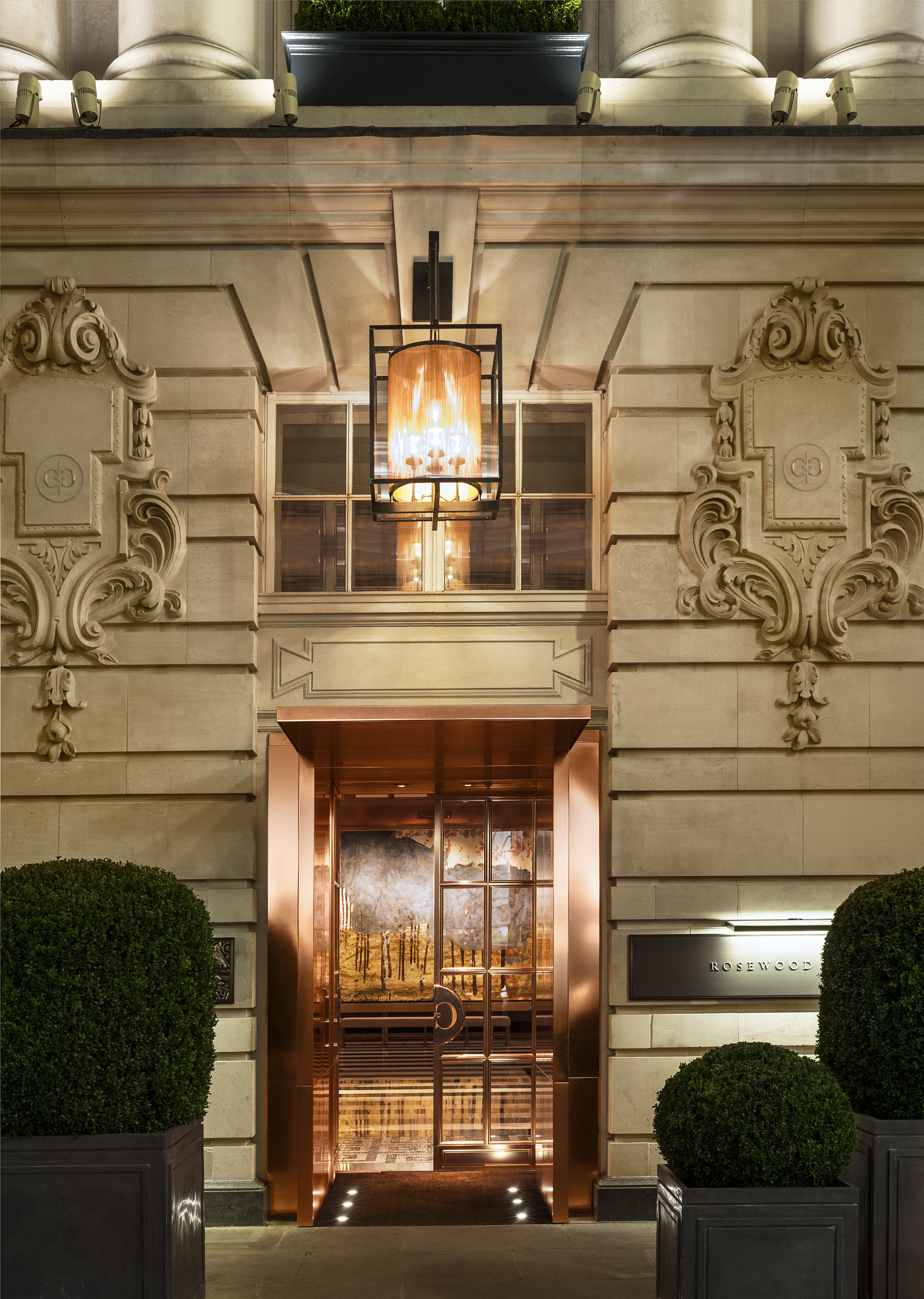 The copper-clad entrance doors of Rosewood London lit at night beneath a hanging lantern, framed by carved Edwardian stonework and clipped box topiary in planters