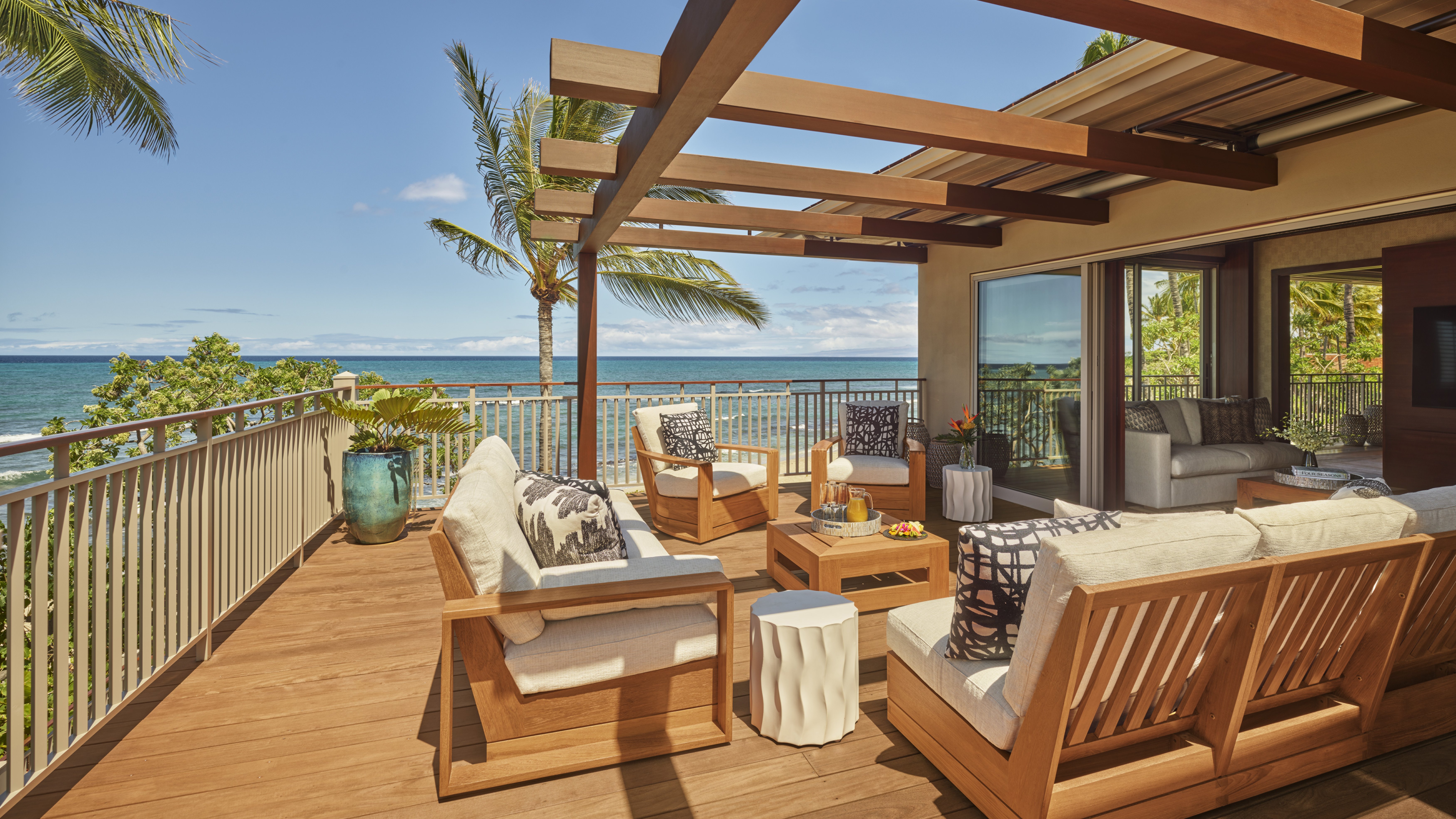 An oceanfront lanai at Four Seasons Resort Hualalai with teak sofas and armchairs under a timber pergola, a potted palm, and the Pacific beyond the balcony rail