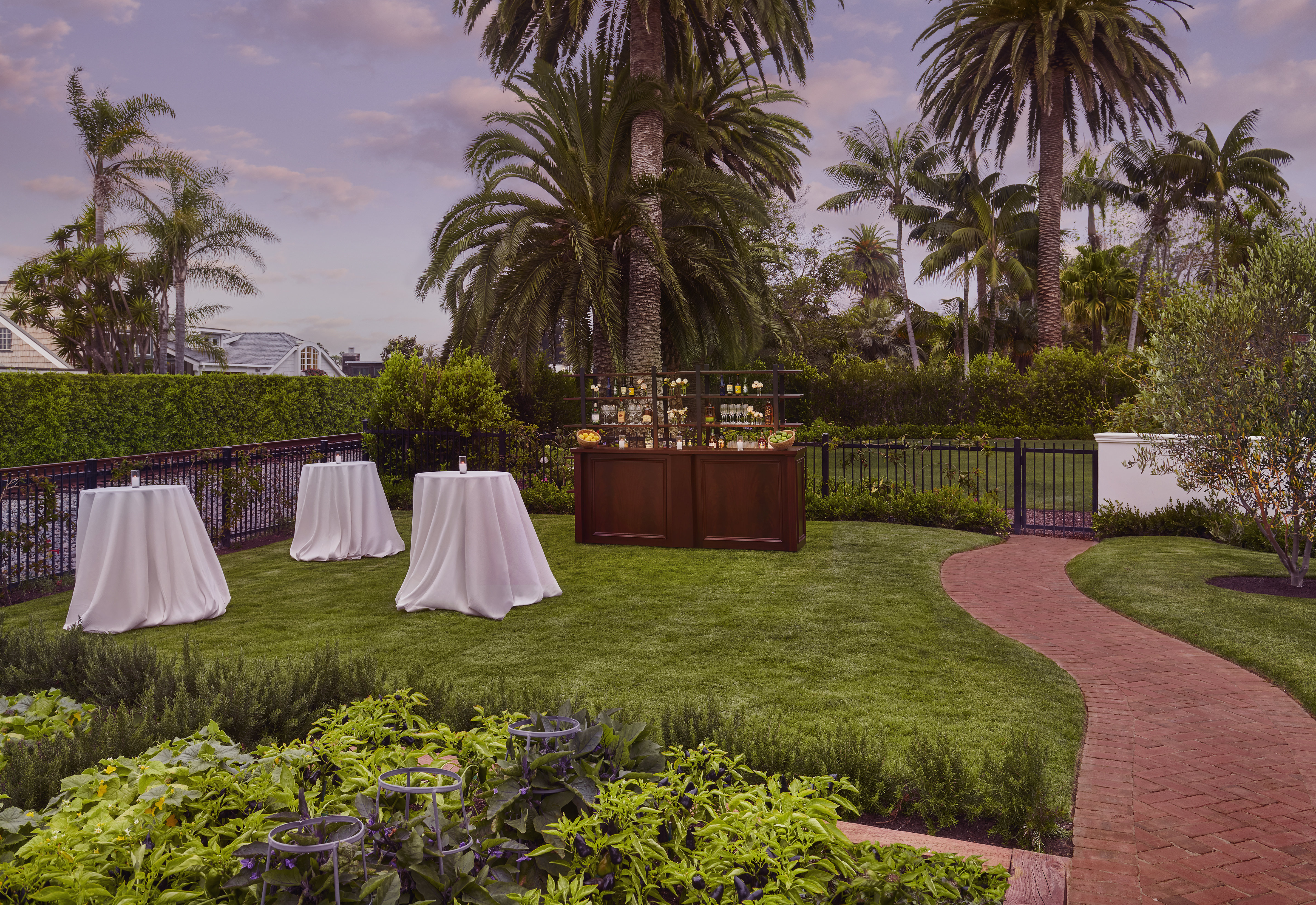 A garden lawn at Rosewood Miramar Beach set for an evening reception with draped cocktail tables and a mahogany bar beneath palms - the kind of unrequested, staged detail that anticipatory 5-star service produces