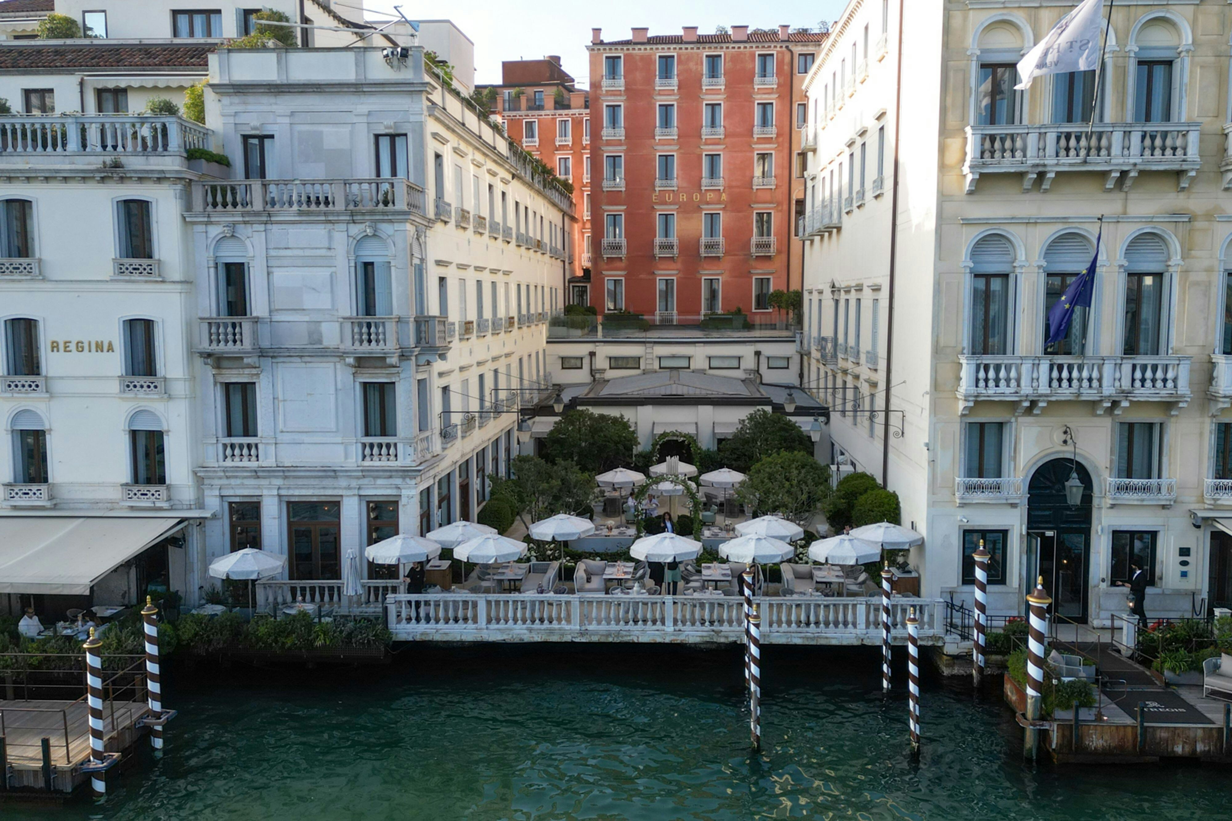 The canal-side garden terrace of The St. Regis Venice with white umbrellas between the Regina and Europa palazzi on the Grand Canal - Venice collects a per-person city tax at the desk that never appears in the online rate