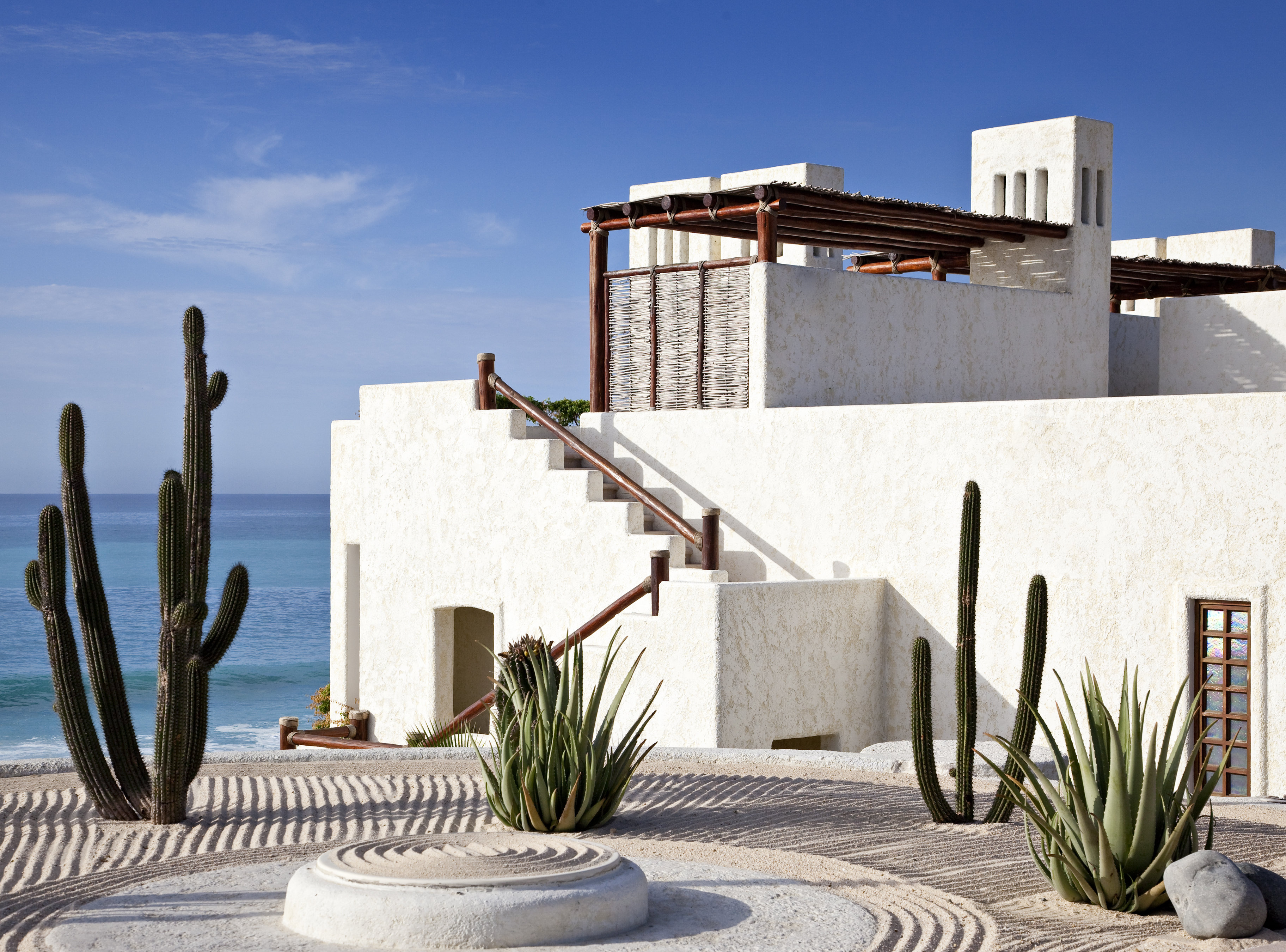 A whitewashed stucco suite building at Las Ventanas al Paraiso with an external staircase rising to a pergola-shaded rooftop terrace, cardon cacti and aloes in raked sand, and the Sea of Cortez beyond