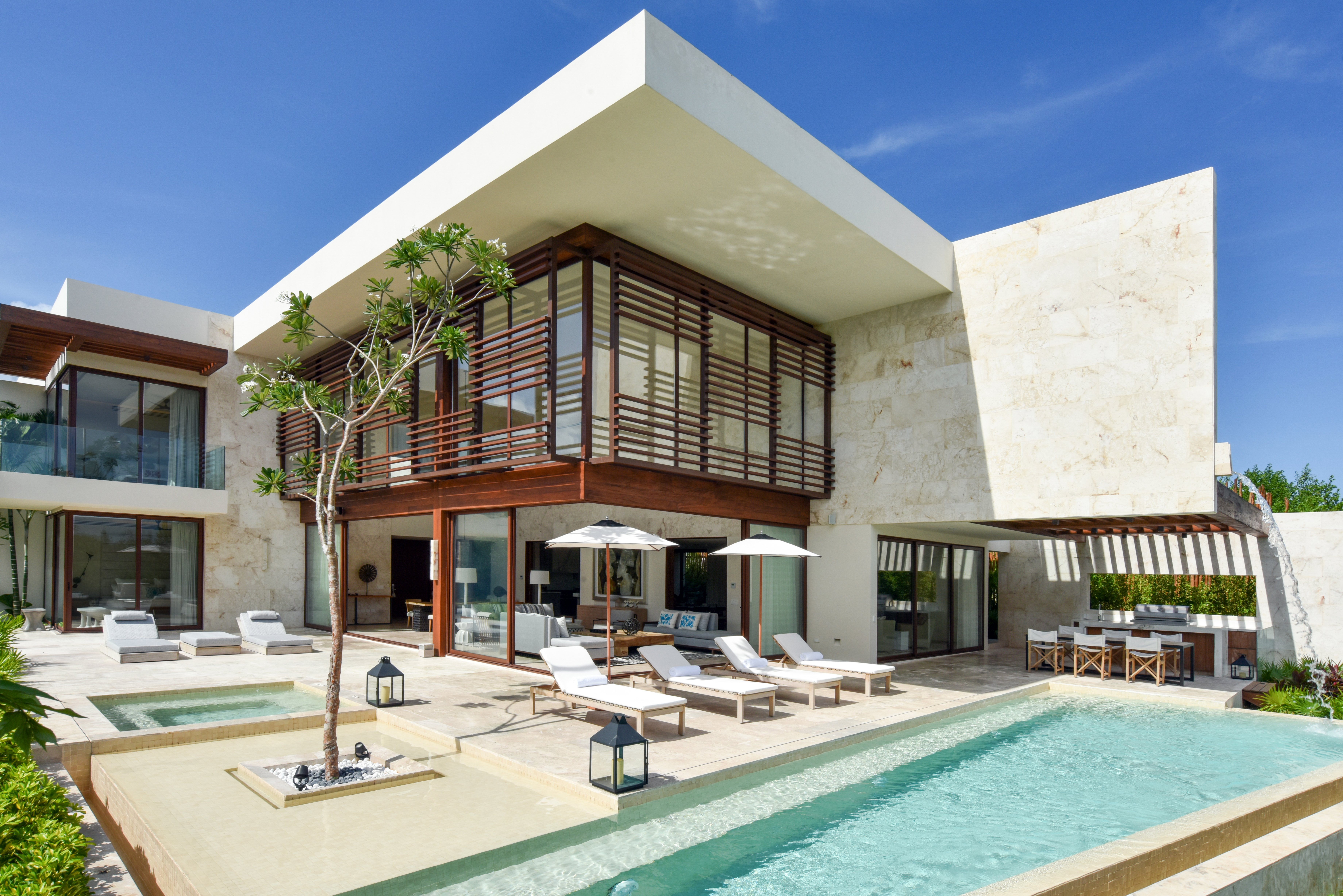 A two-storey villa at Rosewood Mayakoba with pale stone walls, timber-slatted upper floor and wide glass doors opening onto a sun terrace with white loungers, parasols, an outdoor dining table and a long private pool with a plunge pool and a frangipani tree