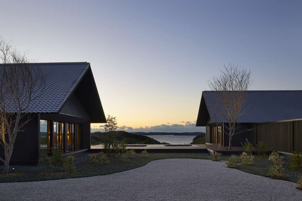 Two dark-tiled pavilions with steep pitched roofs at Amanemu in Ise-Shima, Japan, framing a still hot-spring pool and a view over Ago Bay at dusk, with a raked gravel path and bare trees in the foreground