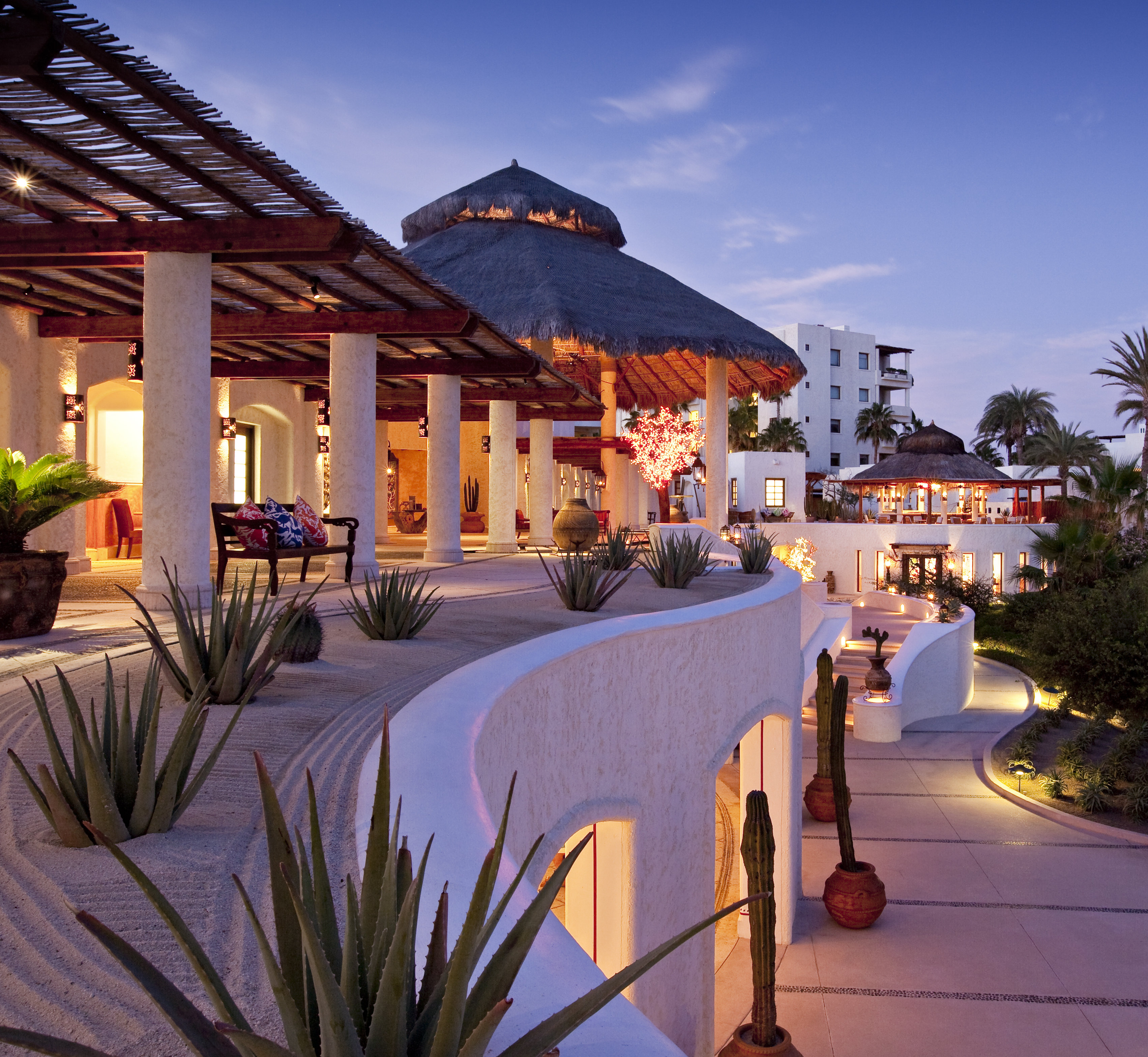 The arrival terrace at Las Ventanas al Paraiso at dusk: a colonnade under a reed pergola, a bench with embroidered cushions, aloes in raked sand, a thatched palapa roof and lantern-lit paths curving down the whitewashed walls