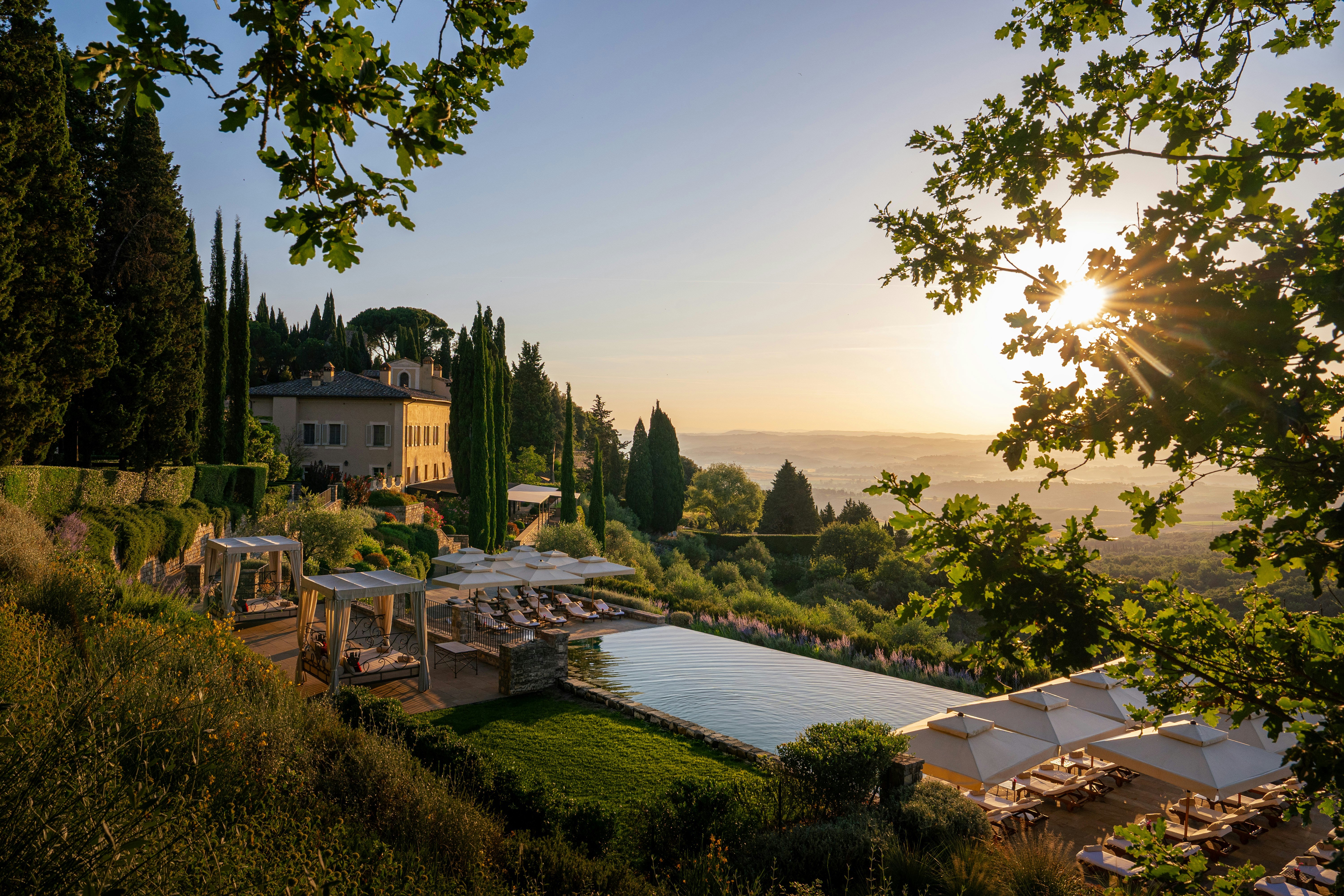 Sunrise over the Val d'Orcia at Rosewood Castiglion del Bosco, with a long infinity pool, canopied day beds and parasols on a terrace below the estate's stone villa and cypress trees