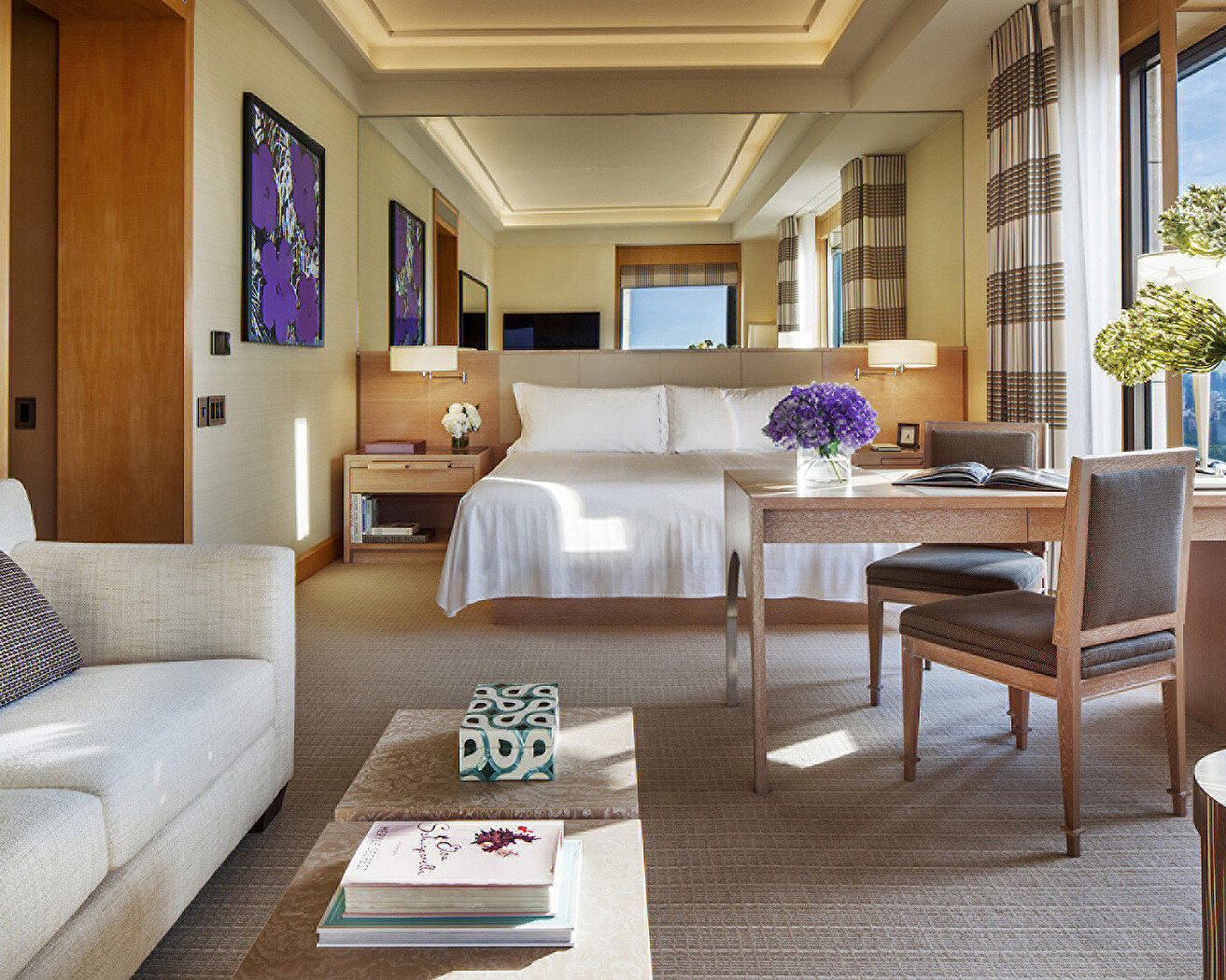 A king room at Four Seasons Hotel New York with a white bed against a pale timber headboard, a sofa and marble coffee table, a writing desk with a vase of purple hydrangeas, Warhol-style flower prints on the wall and tall windows with striped curtains