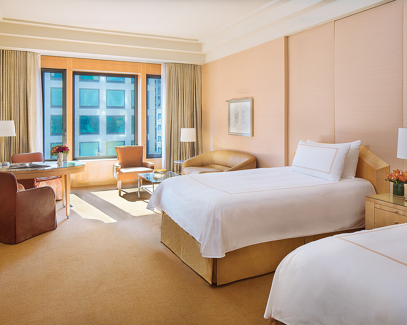 A twin-bedded guest room at Four Seasons Hotel New York with pale sycamore panelling, white bed linen, an armchair and writing desk by the window and a view of the building opposite