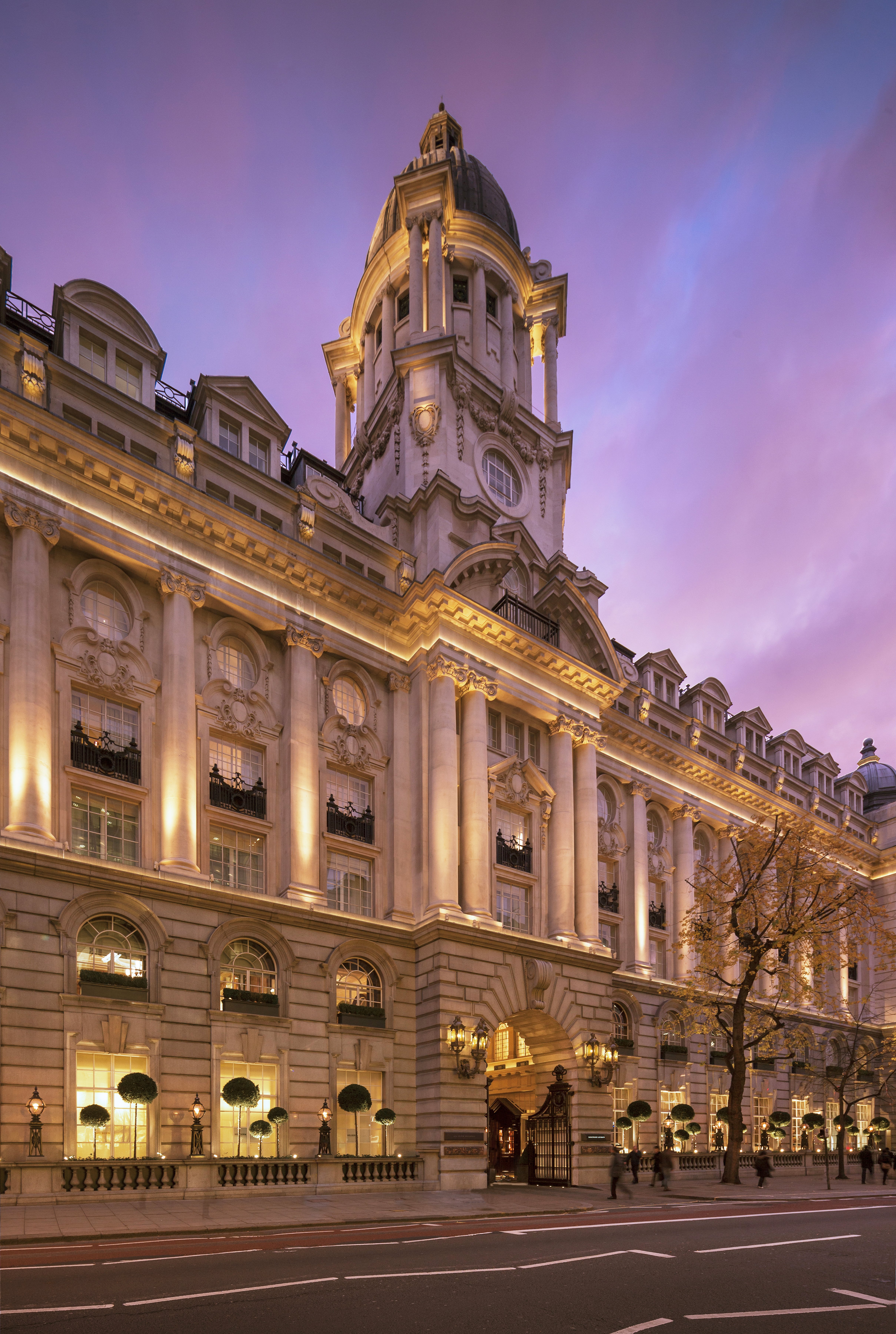 The Edwardian facade of Rosewood London on High Holborn lit at dusk under a violet sky, with a domed corner tower, arched carriage entrance with lanterns and clipped topiary along the street frontage