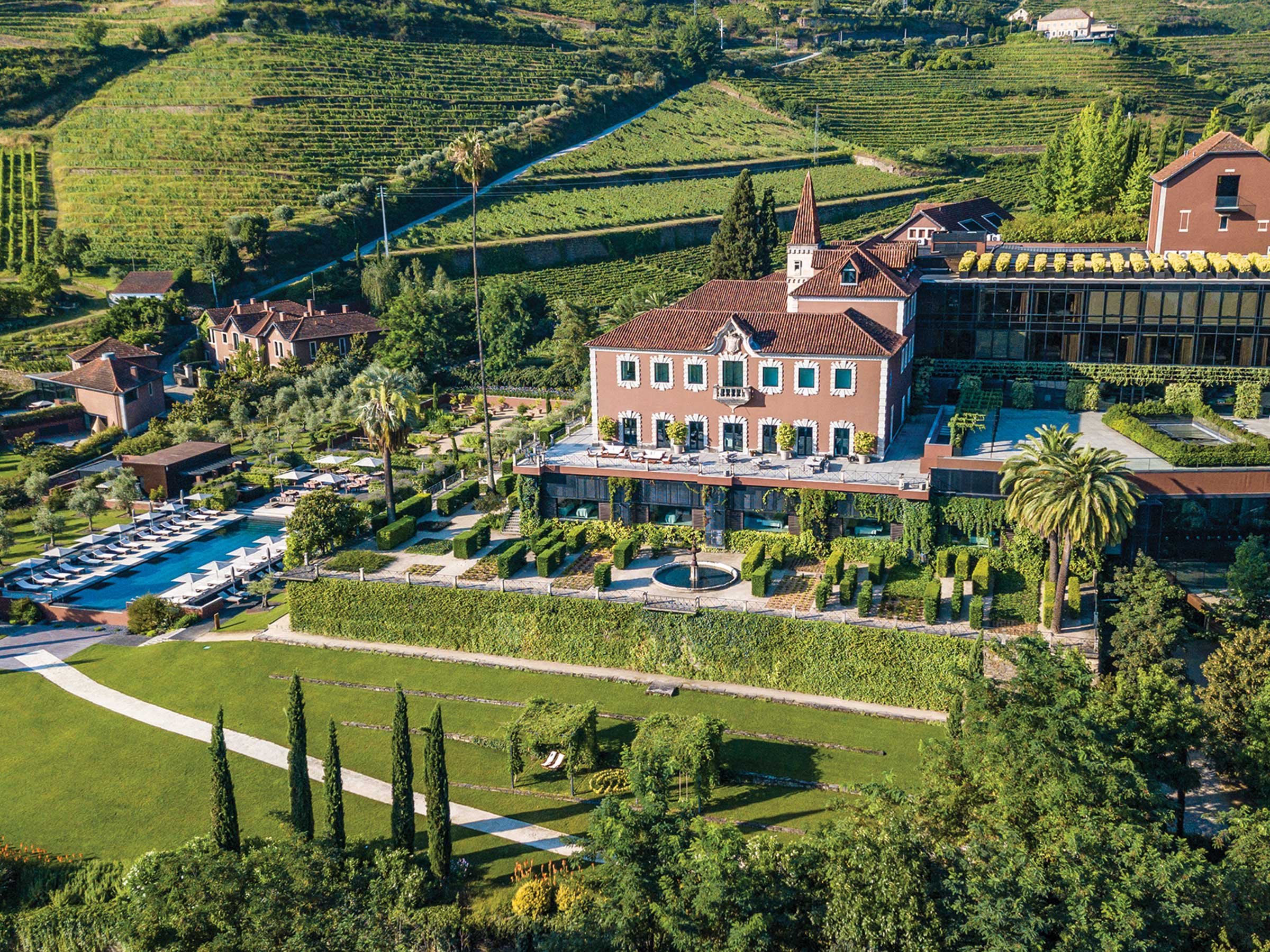Aerial view of Six Senses Douro Valley in Portugal - a 19th-century manor house with terracotta roofs set among terraced vineyards, with a long outdoor pool lined with loungers and parasols, formal box-hedge gardens, cypresses and a glass-fronted spa wing built into the hillside