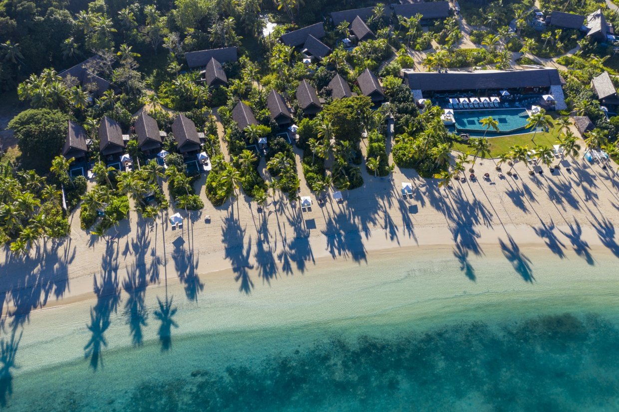Aerial view of the Six Senses Fiji beachfront villa line with the substantial thatched-roof pool villas set behind the palms of the western Malolo Island shore and the dedicated reef-sheltered lagoon shallows