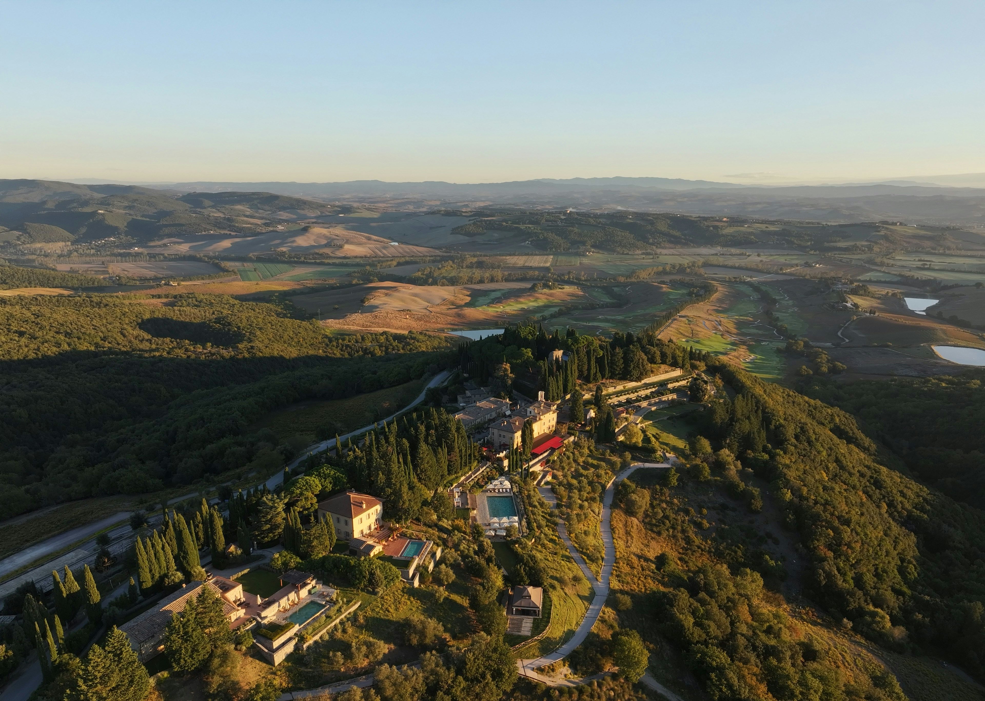 Aerial view at golden hour of Rosewood Castiglion del Bosco on a Tuscan hilltop, the stone borgo and villa among cypresses with several private swimming pools, a main pool terrace and the rolling Val d'Orcia and golf course beyond