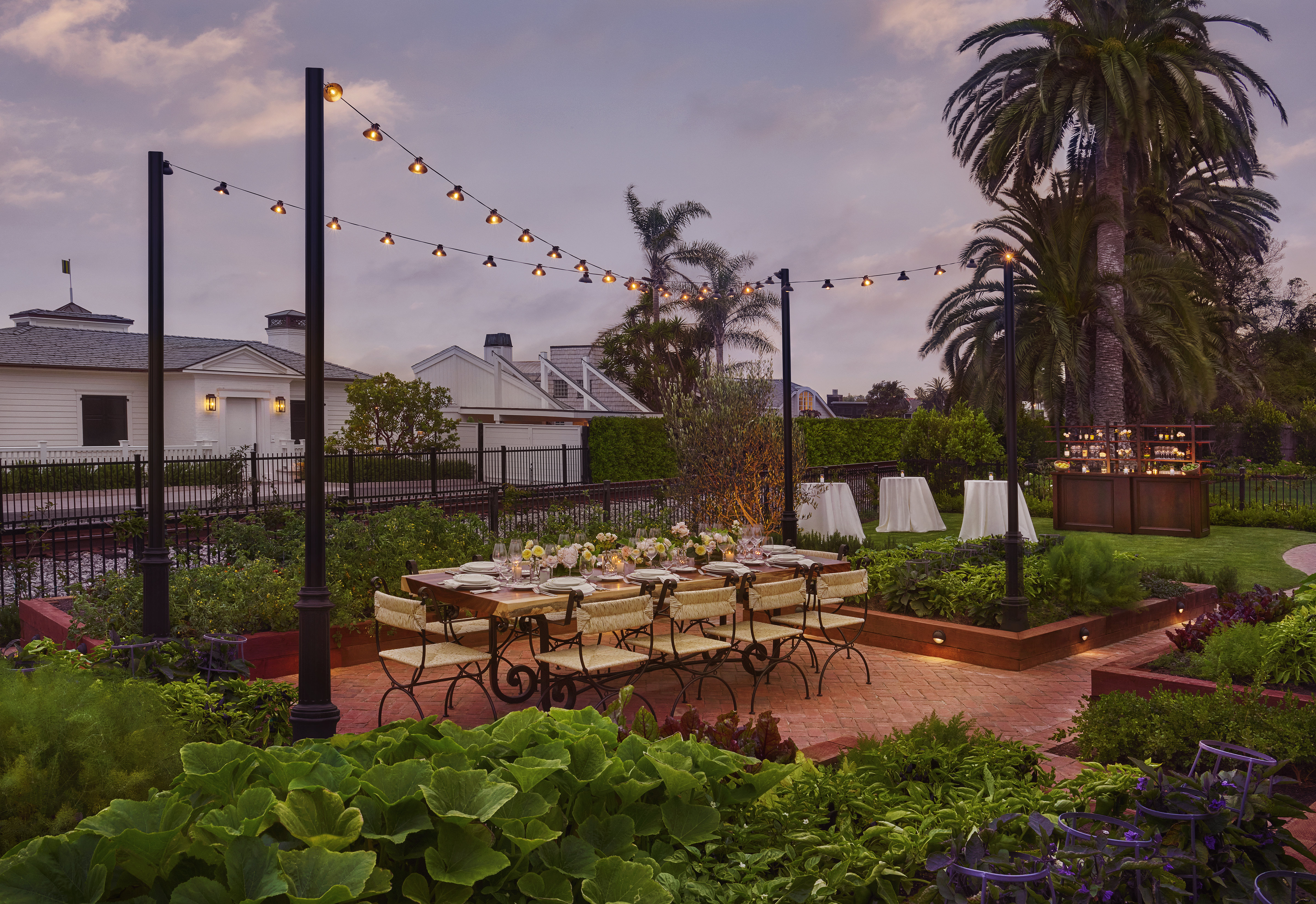 A long table set for dinner at dusk in the kitchen garden at Rosewood Miramar Beach, with festoon lighting strung overhead, raised planting beds and a bar station beyond - the culinary infrastructure a 5-star property maintains beyond its restaurants