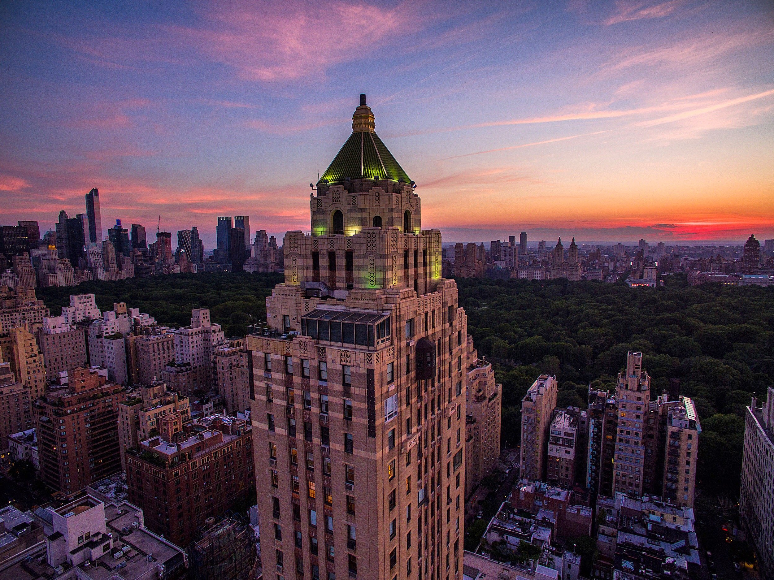 The Art Deco tower of The Carlyle, A Rosewood Hotel, with its lit green pyramidal crown rising above Manhattan's Upper East Side at sunset, Central Park and the Midtown skyline behind