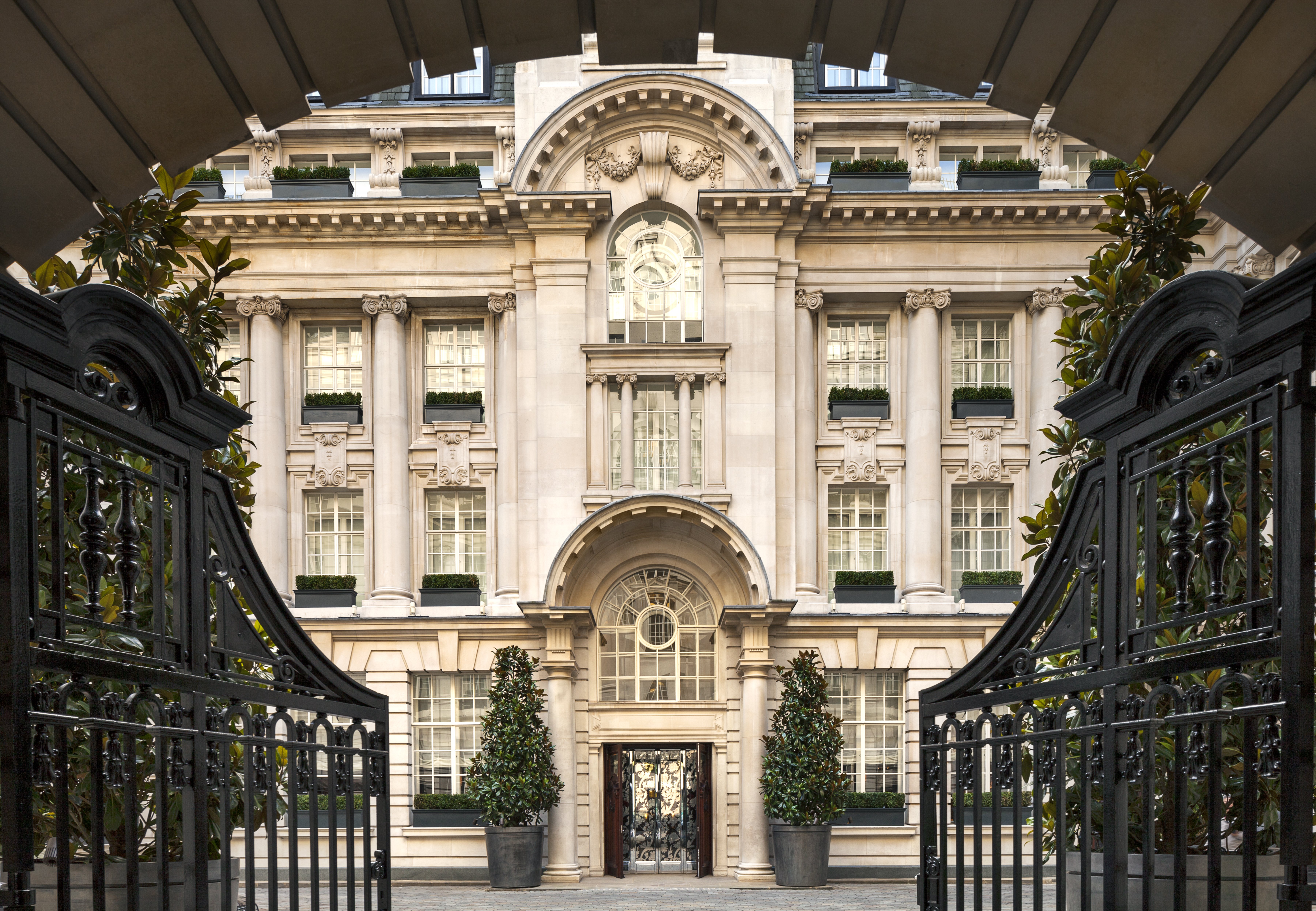 The Edwardian stone courtyard facade of Rosewood London viewed through the arched carriage entrance, with wrought-iron gates, magnolia topiary in planters and window boxes on every floor