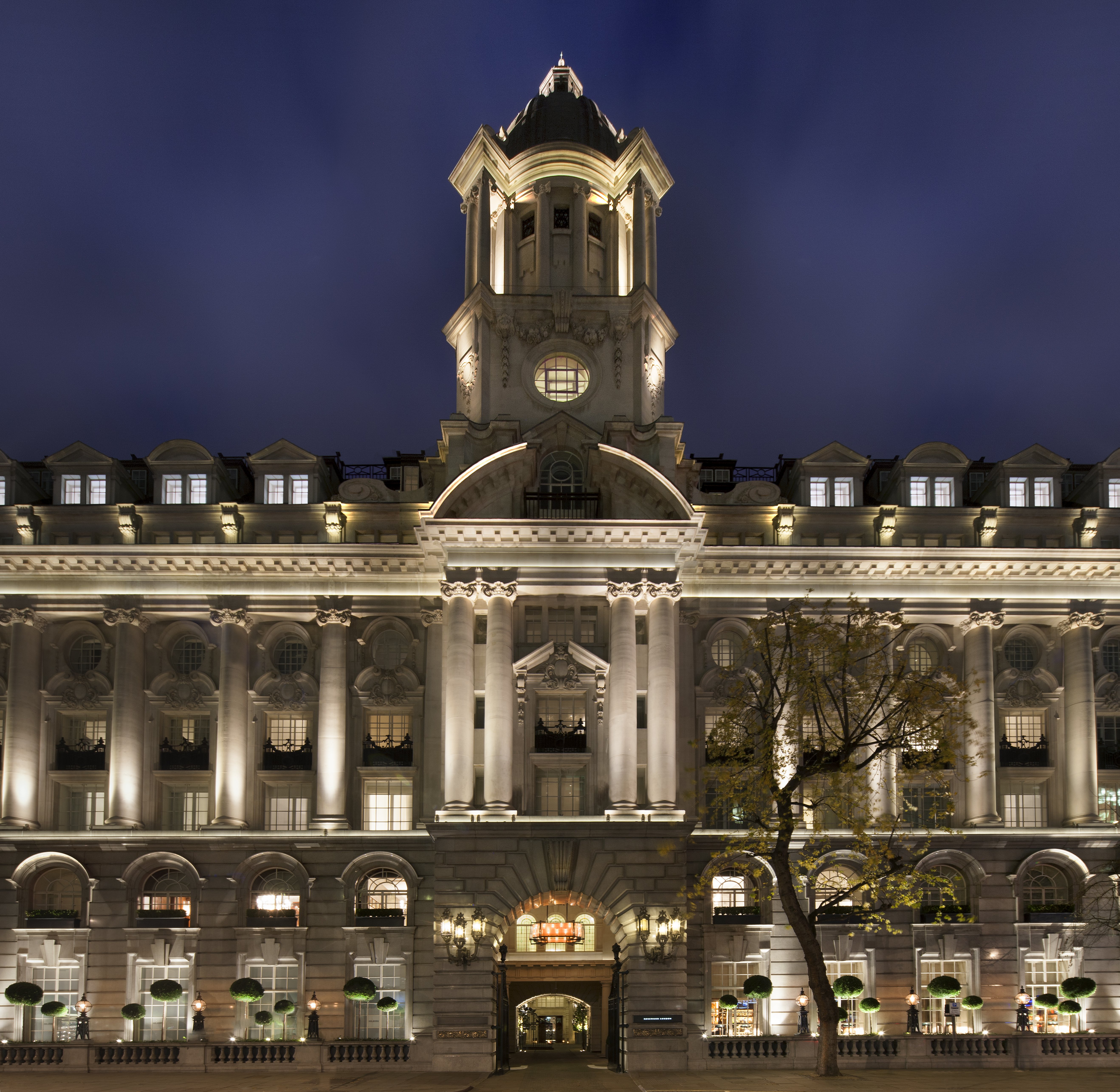 The floodlit Edwardian stone facade and domed clock tower of Rosewood London on High Holborn at night, with its columned upper storeys, lantern-lit carriage archway entrance and clipped bay trees along the pavement