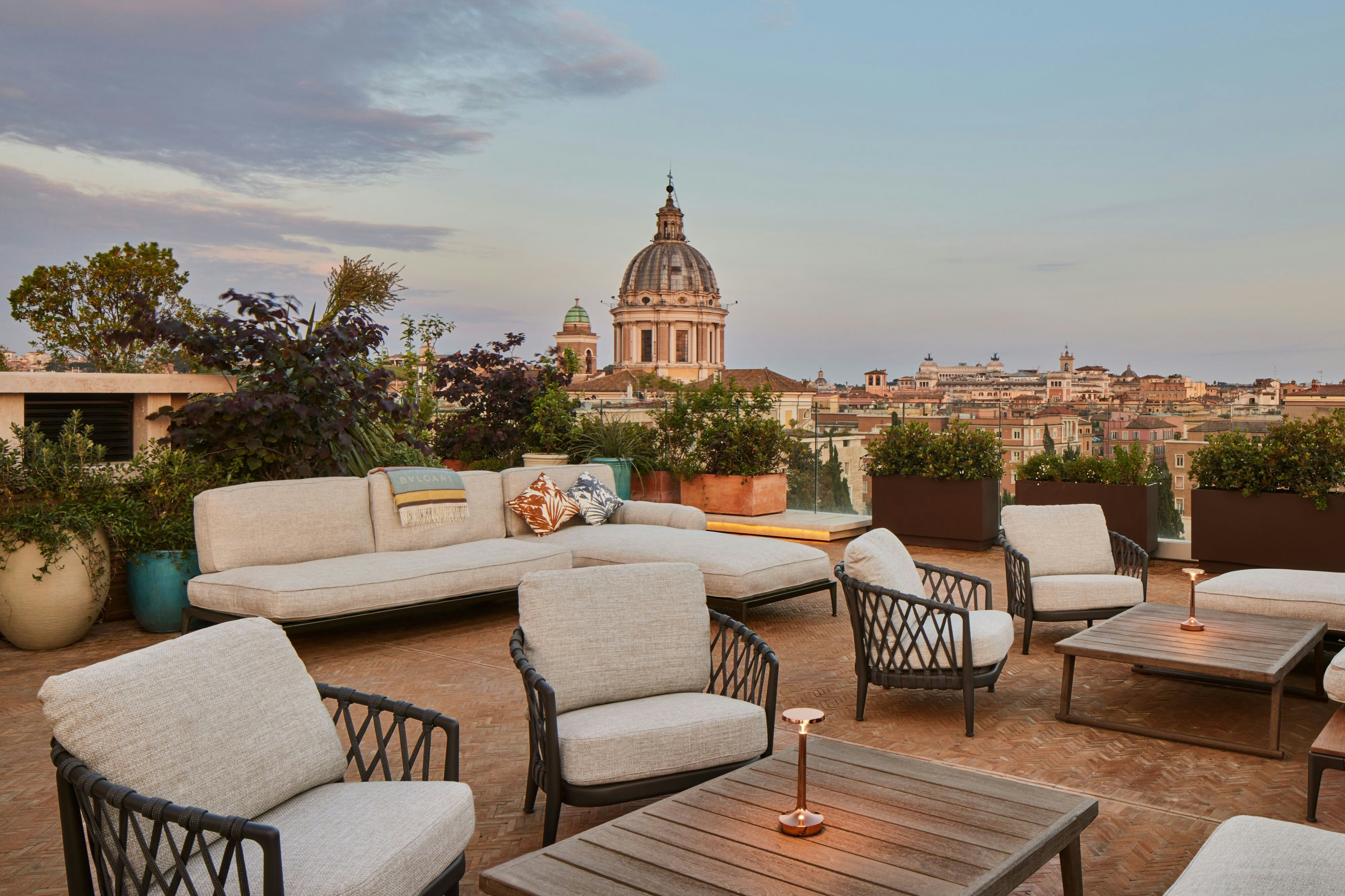 The rooftop terrace at Bulgari Hotel Roma at dusk, low sofas and armchairs around timber tables with copper lamps lit, planters framing a view over Roman rooftops to the dome of San Carlo al Corso
