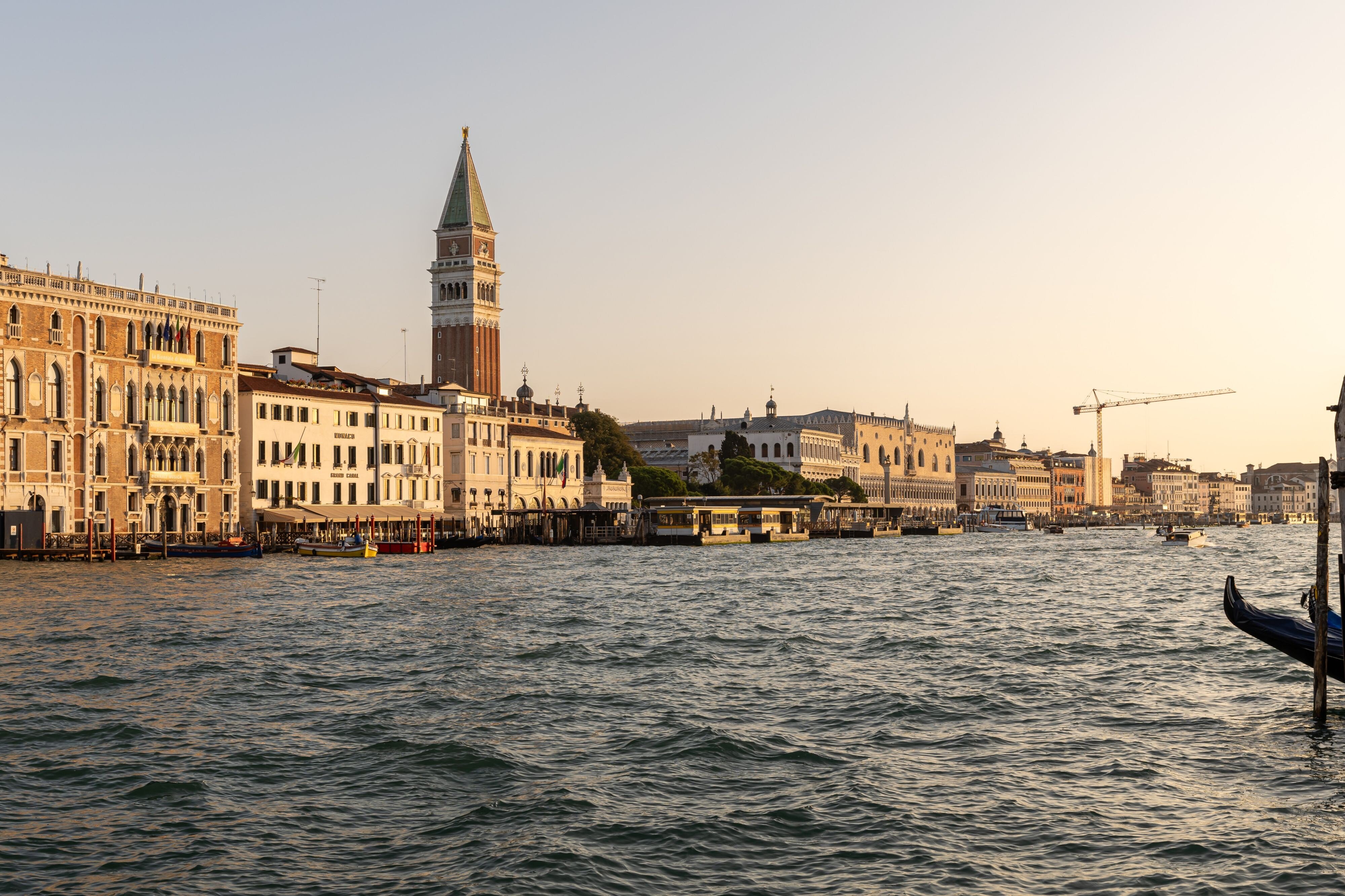 The Venice lagoon at sunset seen from the water, with the campanile of St Mark's, the Doge's Palace and the line of palazzi along the waterfront in warm light, a gondola prow in the foreground